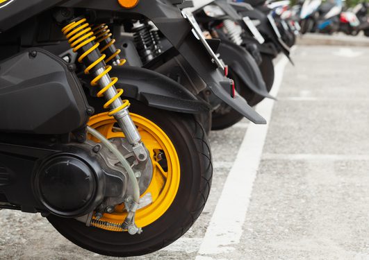 Close-up of a black motorcycle rear wheel with bright yellow suspension springs and yellow rim, parked among other bikes in a paved car park.