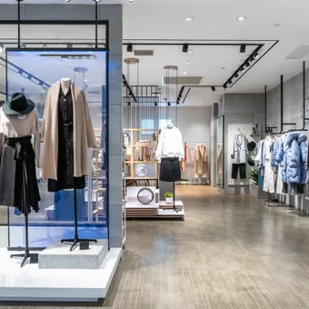 Modern clothing store with a glass-front display on the left showing mannequins in beige and black outfits; coats line the right wall.