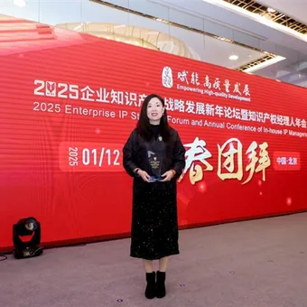 A woman standing on stage in front of a large red backdrop at a conference, dressed in black with a mask, holding an award or certificate.