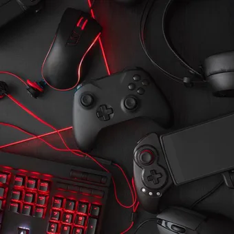 Dark desk with red LED gaming gear: backlit keyboard, glowing mouse, two game controllers, headset and a handheld gaming device.