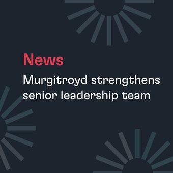 Head-and-shoulders portrait of a person with light blond hair, wearing a dark suit and teal tie, smiling softly against a plain grey background.