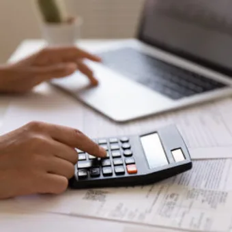 Hands using a calculator on a desk with a laptop and papers in the background.