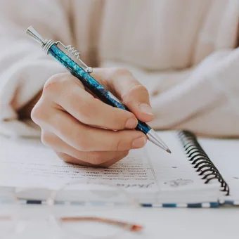 Close-up of a hand holding a blue patterned pen and writing in a spiral notebook on a white table.
